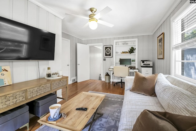 living room with ceiling fan, a decorative wall, wood finished floors, visible vents, and ornamental molding
