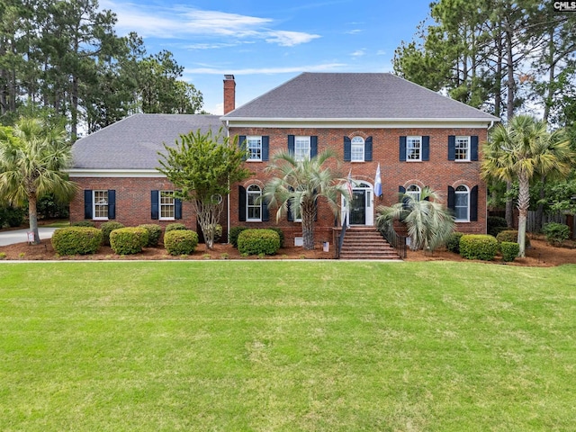 colonial home featuring roof with shingles, a chimney, brick siding, and a front yard