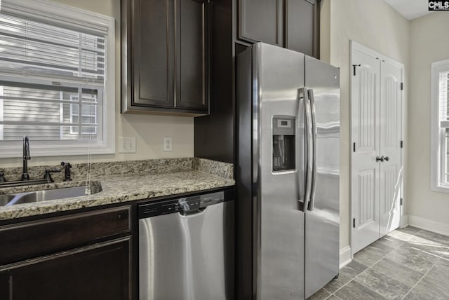 kitchen with appliances with stainless steel finishes, dark brown cabinets, a sink, baseboards, and light stone counters