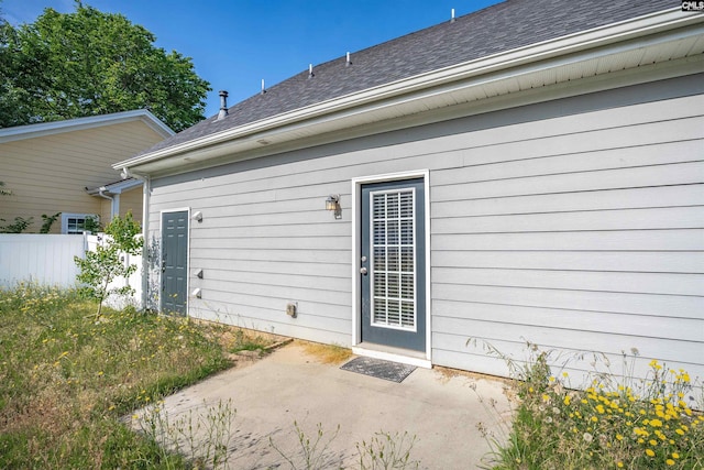 view of exterior entry featuring a patio, a shingled roof, and fence