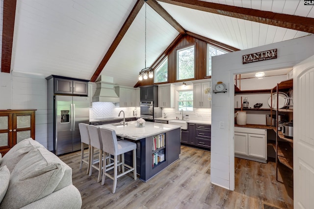 kitchen featuring appliances with stainless steel finishes, a sink, light wood finished floors, vaulted ceiling with beams, and custom range hood