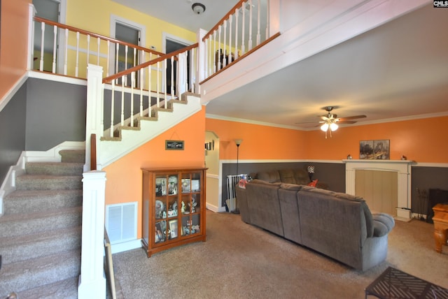 carpeted living room featuring a ceiling fan, visible vents, crown molding, baseboards, and stairs