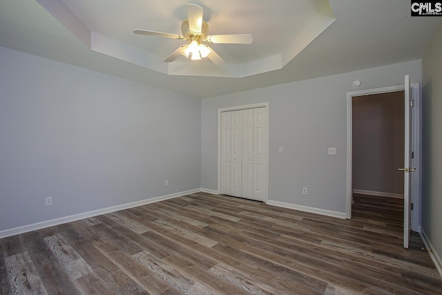 unfurnished bedroom with a closet, dark wood-style flooring, baseboards, ceiling fan, and a tray ceiling