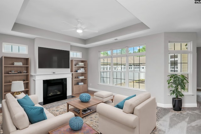 living area featuring a fireplace with flush hearth, a raised ceiling, baseboards, and a ceiling fan