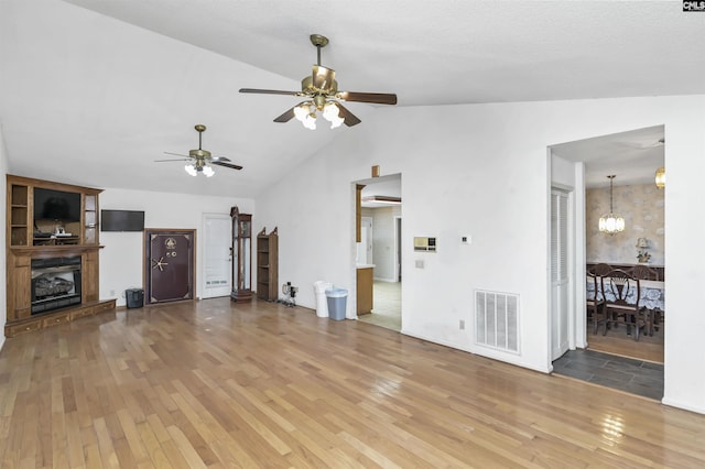 unfurnished living room featuring ceiling fan with notable chandelier, wood finished floors, visible vents, vaulted ceiling, and a glass covered fireplace