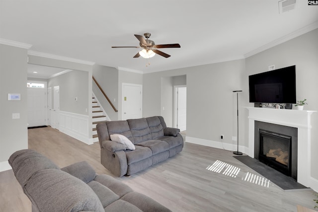 living room featuring wood finished floors, stairs, ceiling fan, a glass covered fireplace, and crown molding