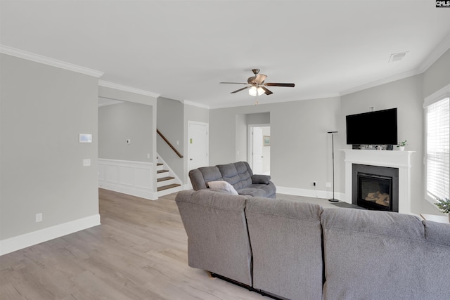 living room with ceiling fan, stairs, light wood-type flooring, a glass covered fireplace, and crown molding