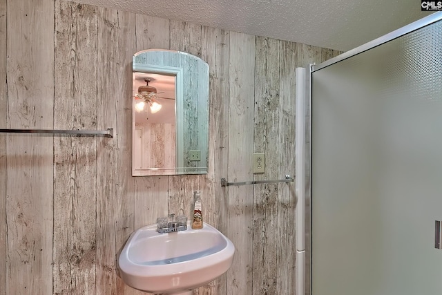 bathroom with wooden walls, a sink, a shower with shower door, a ceiling fan, and a textured ceiling