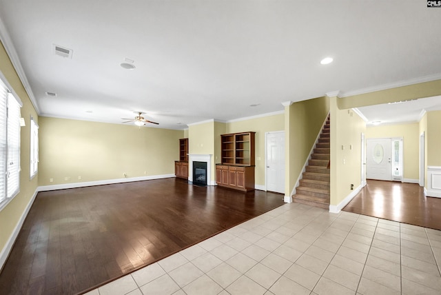 unfurnished living room featuring ceiling fan, stairs, ornamental molding, a glass covered fireplace, and wood finished floors