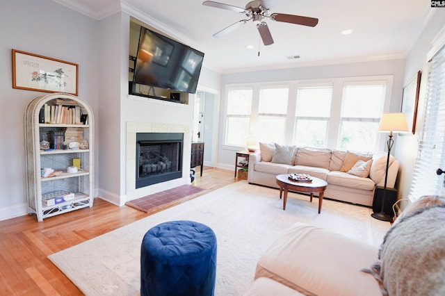 living area with a ceiling fan, wood finished floors, ornamental molding, a tiled fireplace, and baseboards