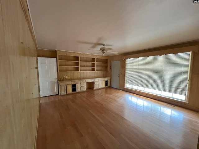 unfurnished living room featuring light wood-style flooring, a ceiling fan, wood walls, built in features, and crown molding