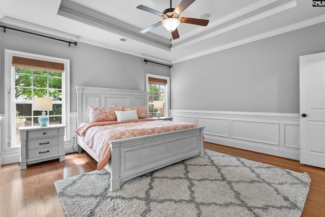 bedroom with crown molding, a raised ceiling, multiple windows, wood finished floors, and a wainscoted wall