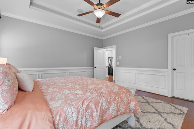 bedroom featuring a raised ceiling, ornamental molding, wood finished floors, wainscoting, and a ceiling fan