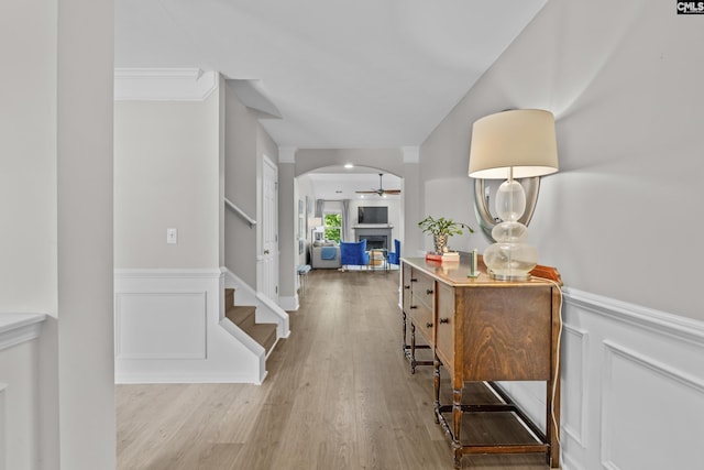 hallway featuring a wainscoted wall, light wood finished floors, arched walkways, stairway, and ornamental molding