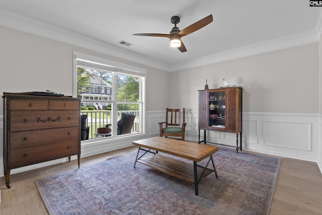 sitting room with a ceiling fan, ornamental molding, wainscoting, wood finished floors, and a decorative wall