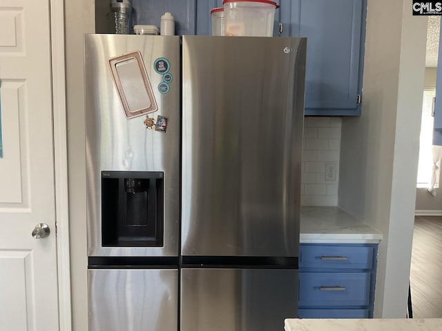 kitchen view of stainless steel fridge, blue cabinets, backsplash, and light stone counters