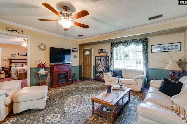 living area featuring ornamental molding, a ceiling fan, a textured ceiling, a glass covered fireplace, and wainscoting