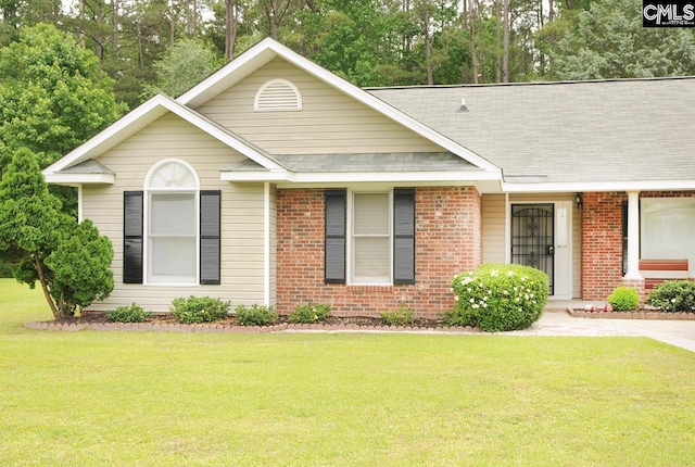 single story home with a front lawn, a shingled roof, brick siding, and view of wooded area
