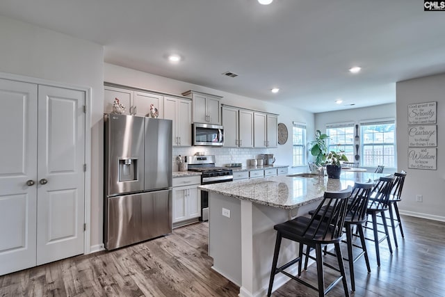 837 Marigold Ln, Lexington, SC 29072 - kitchen featuring appliances with stainless steel finishes, a kitchen island with sink, a breakfast bar area, wood finished floors, and light stone counters