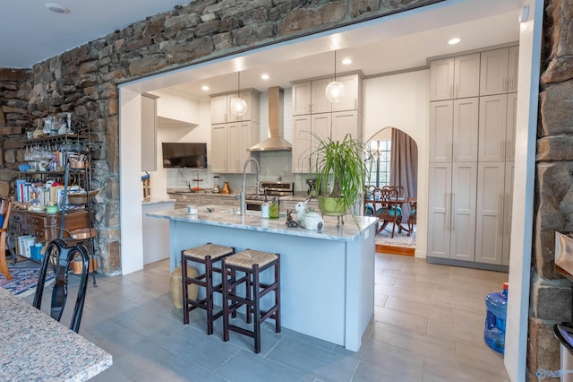 kitchen with decorative backsplash, hanging light fixtures, wall chimney range hood, a breakfast bar, and light stone countertops