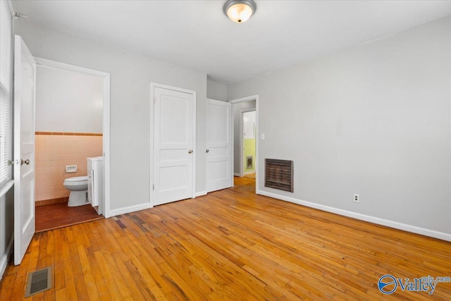 unfurnished bedroom with tile walls, visible vents, ensuite bath, baseboards, and hardwood / wood-style flooring