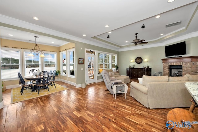 living room with crown molding, a brick fireplace, light hardwood / wood-style flooring, a tray ceiling, and ceiling fan