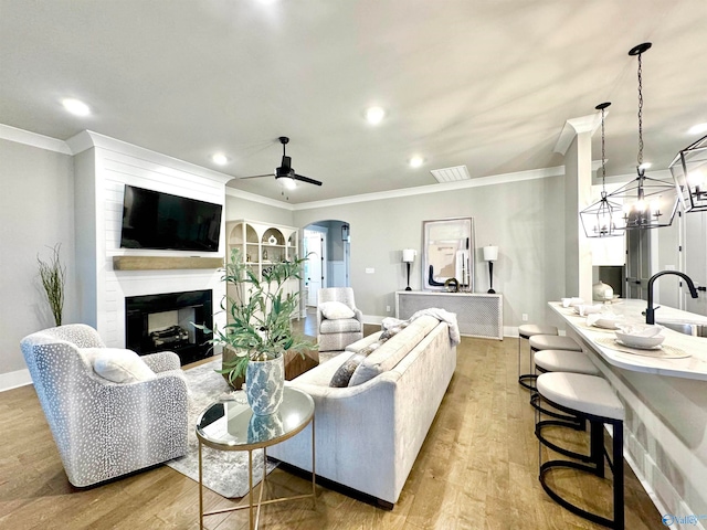 living room with a multi sided fireplace, light hardwood / wood-style flooring, sink, ceiling fan with notable chandelier, and ornamental molding