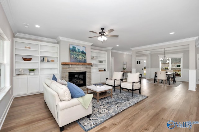 living room featuring built in features, ornamental molding, a fireplace, light wood-type flooring, and ceiling fan with notable chandelier