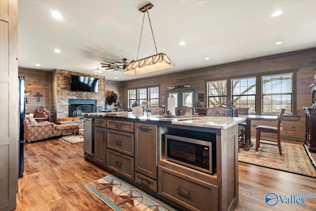 kitchen with a stone fireplace, wooden walls, wood finished floors, open floor plan, and stainless steel microwave