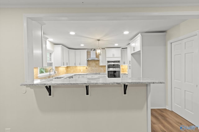 kitchen featuring wall chimney exhaust hood, hanging light fixtures, kitchen peninsula, white cabinetry, and stainless steel double oven