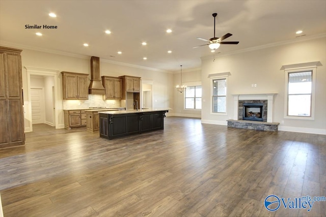 kitchen featuring an island with sink, wall chimney range hood, a fireplace, open floor plan, and ceiling fan with notable chandelier