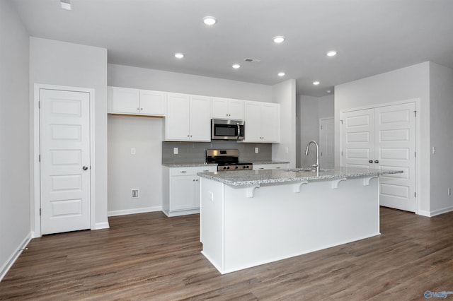 kitchen with dark wood-type flooring, light stone counters, a center island with sink, appliances with stainless steel finishes, and white cabinets