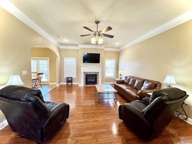 living area with arched walkways, crown molding, a tiled fireplace, wood finished floors, and baseboards