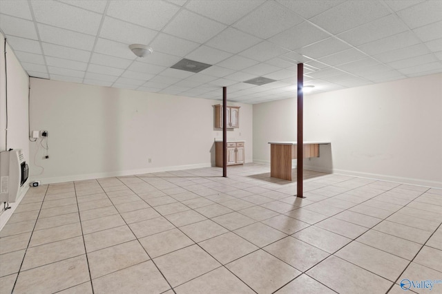 finished below grade area featuring a paneled ceiling, heating unit, light tile patterned flooring, and baseboards
