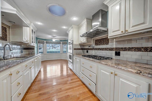 kitchen with light wood-style flooring, a sink, white cabinetry, wall chimney range hood, and appliances with stainless steel finishes