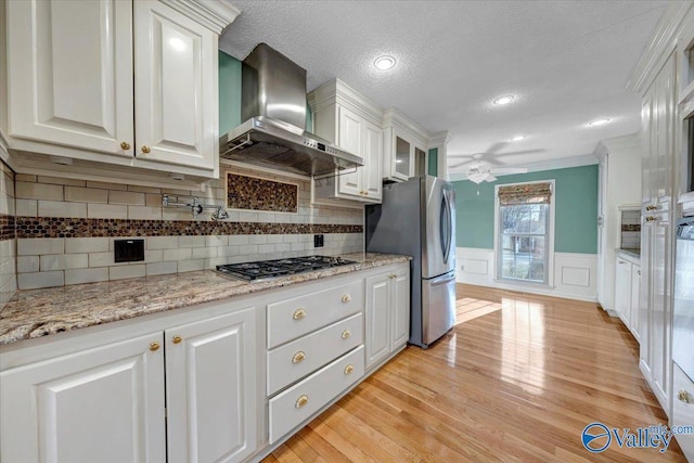 kitchen with light wood finished floors, stainless steel appliances, wainscoting, white cabinets, and wall chimney range hood