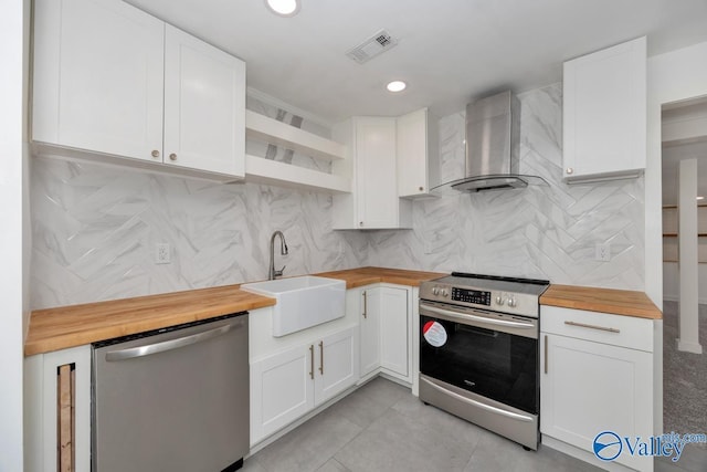 kitchen with open shelves, butcher block counters, wall chimney exhaust hood, a sink, and appliances with stainless steel finishes