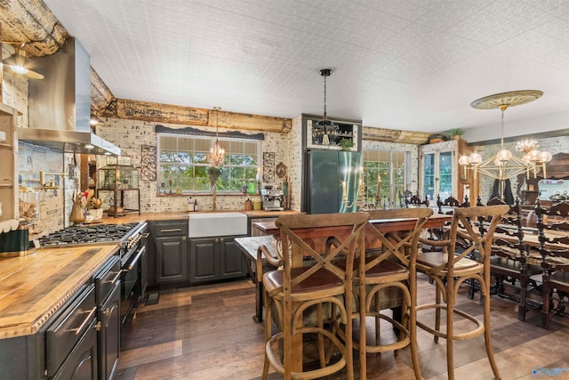 kitchen with sink, a notable chandelier, wall chimney range hood, stainless steel refrigerator, and butcher block countertops