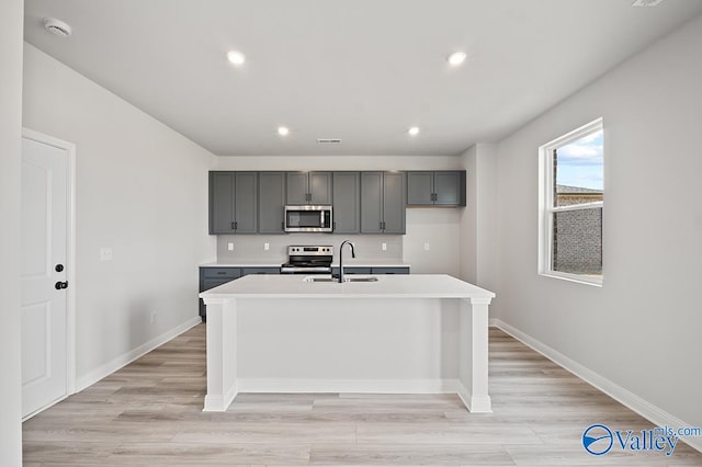 kitchen with gray cabinetry, sink, a kitchen island with sink, appliances with stainless steel finishes, and light wood-type flooring