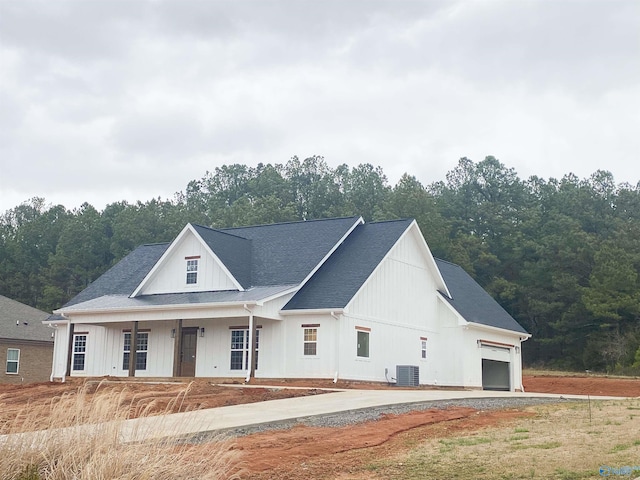 modern inspired farmhouse with board and batten siding, covered porch, a wooded view, a garage, and central AC unit