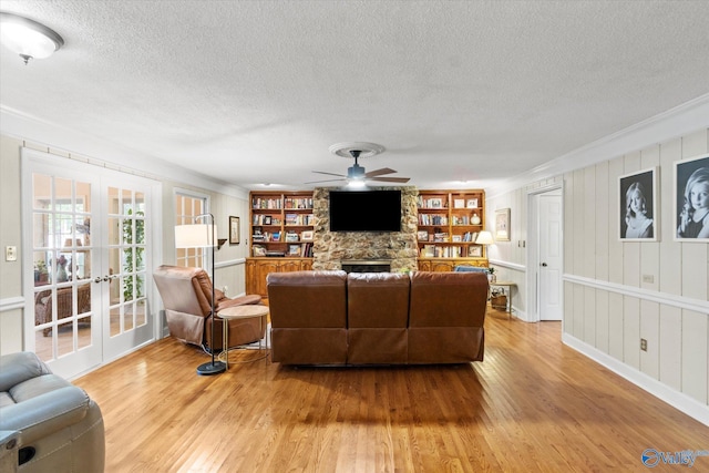 living area with a textured ceiling, a stone fireplace, a ceiling fan, french doors, and light wood-type flooring