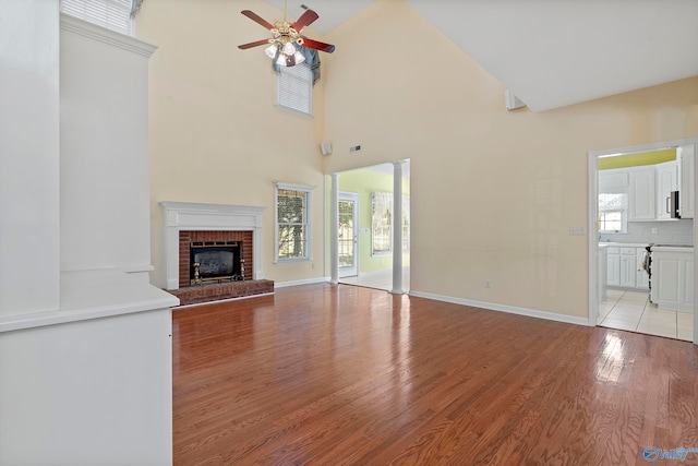 unfurnished living room with light wood-type flooring, ceiling fan, a towering ceiling, and a brick fireplace