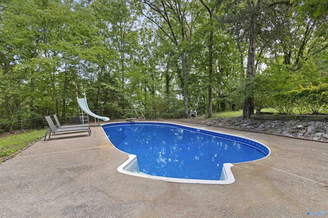 outdoor pool with a water slide, a patio area, a diving board, fence, and view of scattered trees