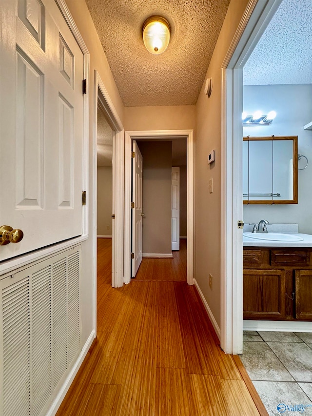 hallway with light wood finished floors, visible vents, a sink, a textured ceiling, and baseboards