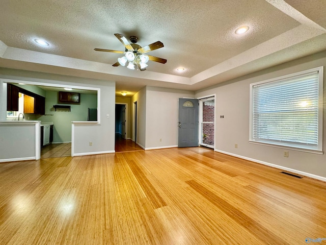 unfurnished living room featuring a raised ceiling, a ceiling fan, a textured ceiling, light wood-type flooring, and baseboards