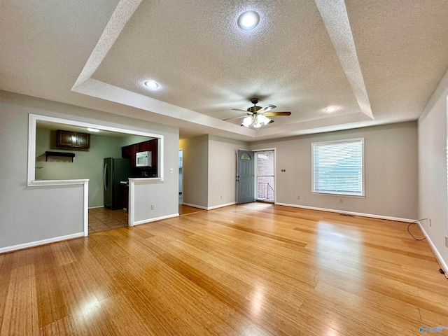 unfurnished living room with a textured ceiling, ceiling fan, a tray ceiling, and light wood-type flooring