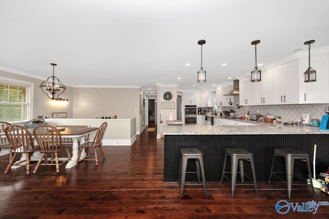 kitchen featuring dark wood finished floors, a peninsula, ornamental molding, white cabinetry, and wall chimney exhaust hood