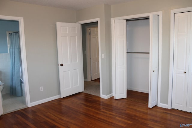 unfurnished bedroom with two closets, baseboards, ensuite bath, and dark wood-style floors