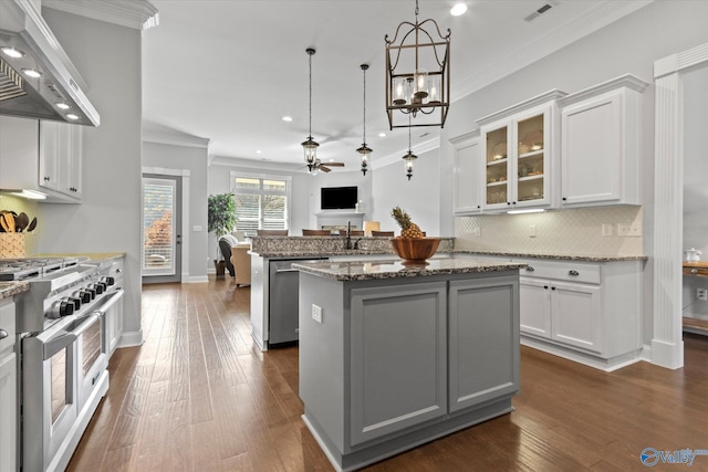 kitchen featuring gas range, wall chimney range hood, an island with sink, white cabinets, and ceiling fan with notable chandelier