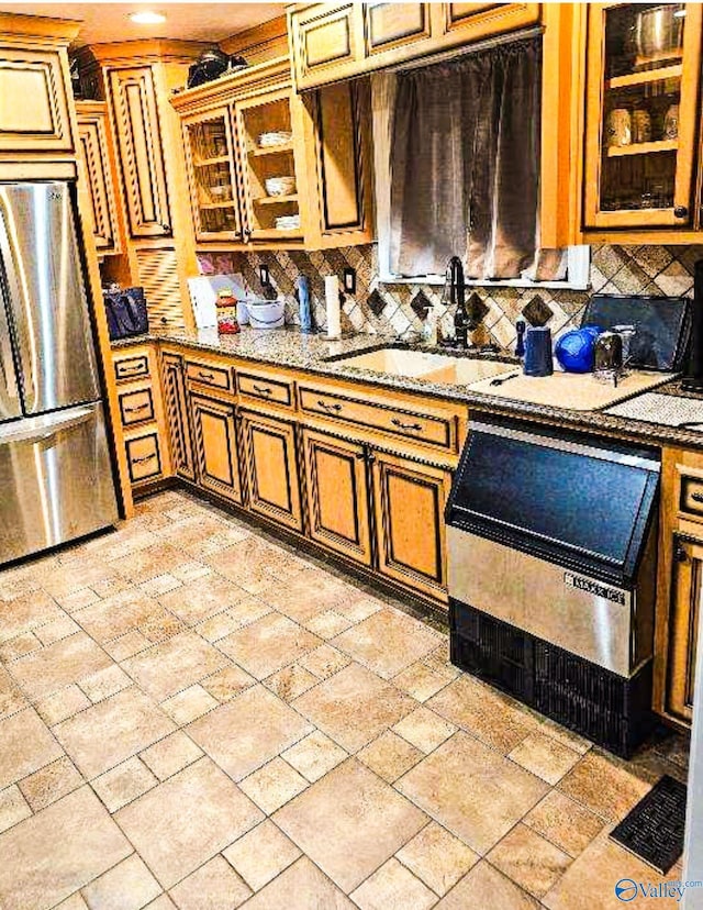 kitchen with a sink, freestanding refrigerator, light stone countertops, decorative backsplash, and glass insert cabinets